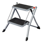 Taburete plegable de acero Habitex de 2 peldaños con plataformas antideslizantes negras y patas con tacos.