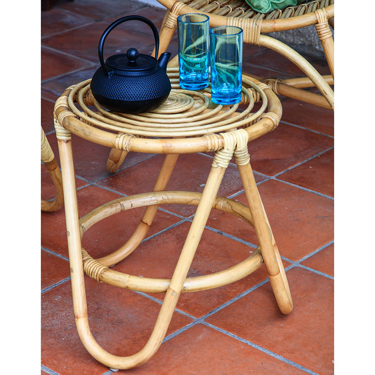 Mesa de centro pequeña Arie de ratán natural ambientada en terraza con tetera y vasos sobre suelo de barro.