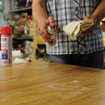 Aplicación de spray limpiador de adhesivos TESA sobre una mesa de madera para quitar manchas de pegamento.