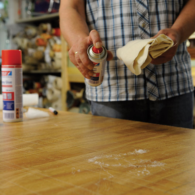Aplicación de spray limpiador de adhesivos TESA sobre una mesa de madera para quitar manchas de pegamento.