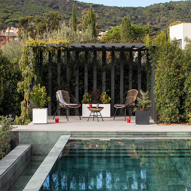 Zona de descanso exterior con conjunto Bistro de ratán junto a una piscina moderna y jardín perimetral.