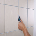 Persona utilizando el rascador Wolfcraft para eliminar mortero en juntas verticales de azulejos beige.
