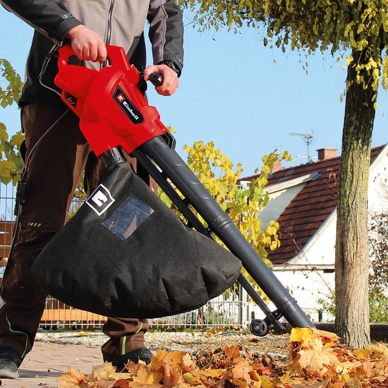Hombre utilizando el aspirador de hojas Einhell GC-EL 3024 E para recoger follaje seco en un jardín.