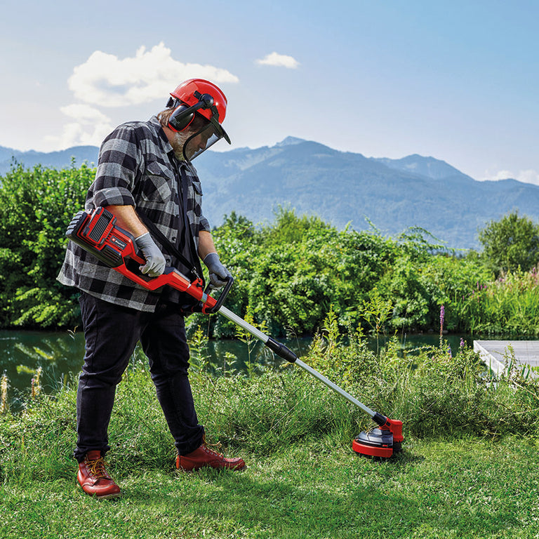 Hombre utilizando la desbrozadora Einhell 36V para cortar hierba alta en un entorno natural cerca de un río.