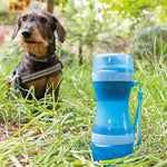 Botella azul 2 en 1 para comida y agua de mascotas situada en el césped con un perro al fondo.