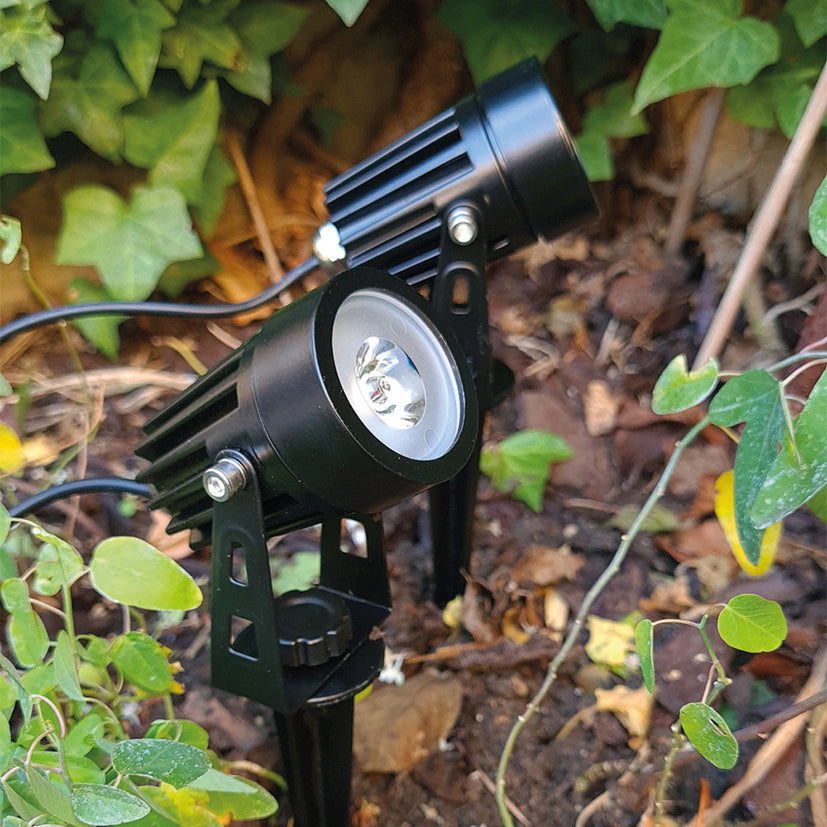 Dos proyectores solares Xanlite instalados con estacas en el suelo de un jardín exterior entre plantas.