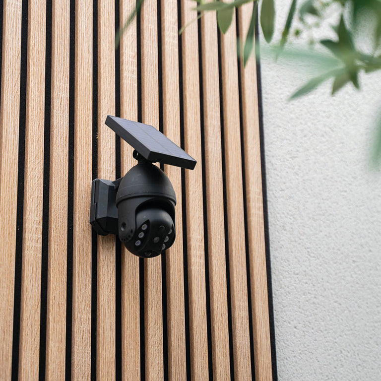 Cámara LED solar Xanlite montada sobre panel de listones de madera decorativo en exterior.