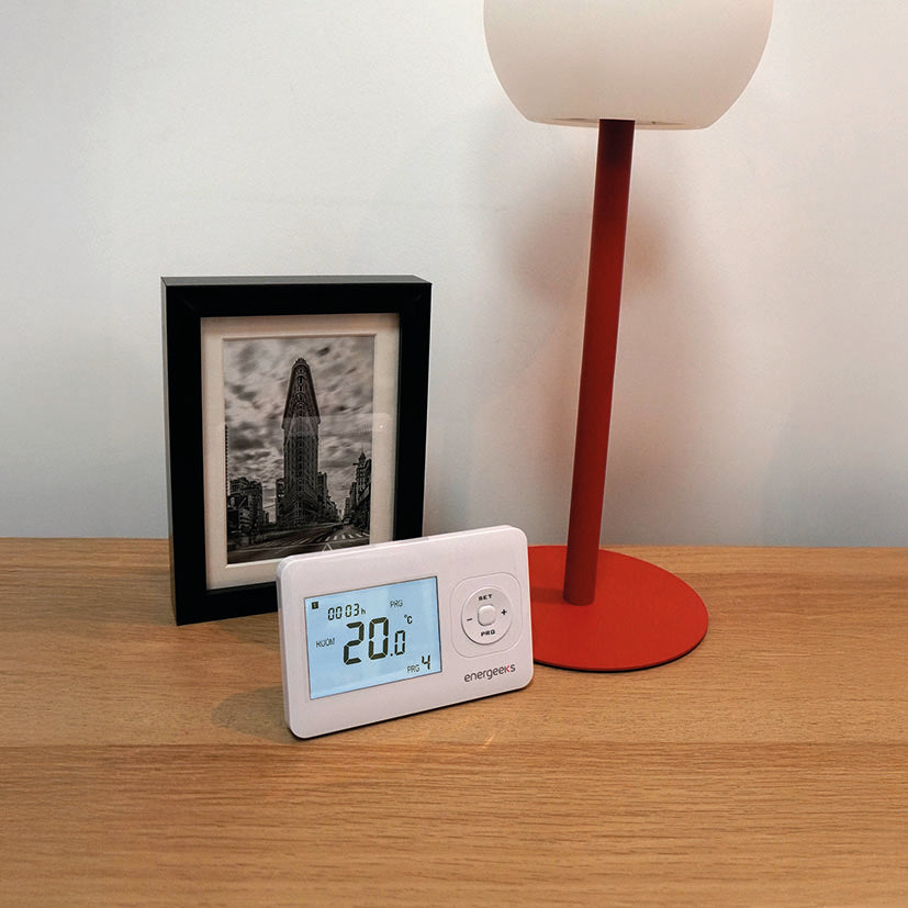 Termostato inalámbrico blanco ENERGEEKS Jasper situado sobre un mueble de madera junto a una lámpara